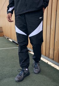 Schwarze Sporthose mit weißen diagonalen Akzenten, aus leichtem Material gefertigt, mit elastischen Bündchen und lockerer Passform. Getragen mit grauen Turnschuhen.