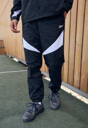 Black athletic pants with white diagonal accents, made of lightweight material, featuring elastic cuffs and a relaxed fit. Worn with grey sneakers.