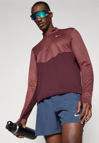 Hombre con camiseta de manga larga color burdeos y rojo oscuro de Nike, pantalones cortos azules de Nike, gafas de sol reflectantes azules, sosteniendo una botella de agua negra.