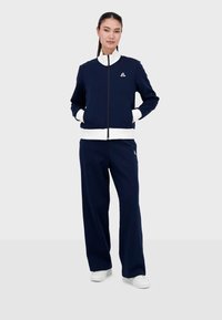 Chándal de color navy con acentos blancos, que incluye una chaqueta con cremallera y cuello alzado, y pantalones a juego. La tela es suave y texturizada, ideal para el uso diario.