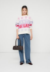 Frau in einem weißen, rosa und roten gemusterten Pullover, blauen Jeans, burgunderfarbenen Schuhen, die eine dunkelbraune Handtasche vor einem schlichten weißen Hintergrund hält.