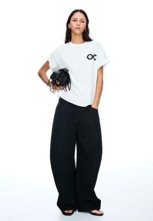 Une mannequin portant un t-shirt blanc ample et un pantalon noir large tient un petit sac à main noir, se tenant devant un fond blanc.