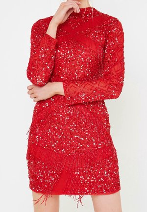 Kobieta ubrana w czerwoną, mini sukienkę z długim rękawem, ozdobioną cekinami i koralikowymi frędzlami w geometryczne wzory na białym tle.