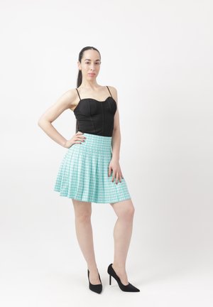 Kvinde med langt mørkt hår iført sort ærmeløs top, lyseblå plisseret nederdel og sorte høje hæle, stående med hånden på hoften.