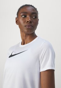 Nike īsām piedurknēm sporta krekls baltā krāsā ar melnu swoosh logotipu uz krūškurvja. Gluda tekstūra, apaļa kakla izgriezums un brīvs piegriezums.