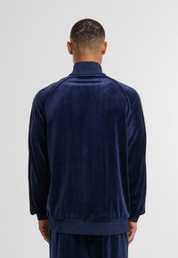 Chaqueta de chándal de terciopelo azul marino con cuello alto, puños acanalados y corte relajado. Textura suave sin patrones visibles ni detalles de hardware.
