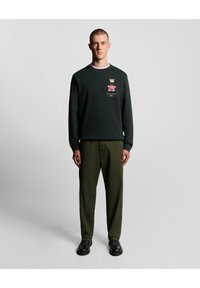 Dunkelgrüner Rundhalsausschnitt-Sweatshirt mit einem Rosensymbol und Krondetail, kombiniert mit olivgrünen Hosen und schwarzen Schnürstiefeln.