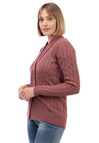 Pletený kardigan v prachově růžové barvě, s kabelovým vzorem, výstřihem do V a žebrovanými manžetami. Nosený přes modré džíny.