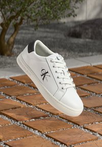 White leather shoe with black heel accent, flat sole, round toe, and laces. Features "Calvin Klein Jeans" logo on the side.