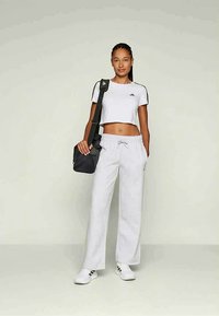 Sieviete, tērpusies baltā crop topā, brīvās baltās sporta biksēs, baltās sporta kurpēs, nēsā melnu pleca somu un stāv uz vienveida fona.