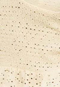 Ύφασμα κρεμ με υφή smocked και μοτίβο από διάτρητα μικρά τετράγωνα, με λεπτομέρεια από μαζεμένο βολάν.