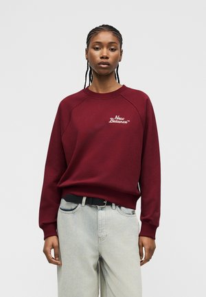 Sieviete ar pīteni, tērpusies bordo krāsas New Balance džemperī un gaiši zilās džinsa biksēs, stāv pret vienkāršu gaiši pelēku fonu.