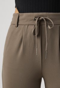 I pantaloni in un morbido tessuto taupe presentano una vita aderente con chiusura a cordoncino, accentuata da aghetti metallici e dettagli plissettati.