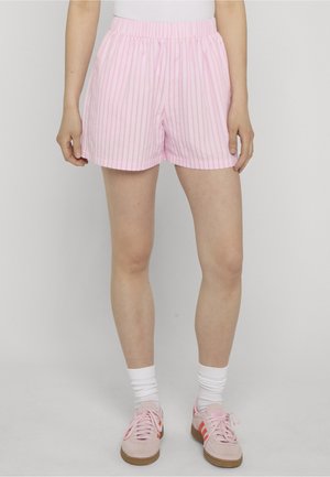 Person trägt pink-weiß gestreifte Shorts, weiße Socken und pinke Sneakers und steht vor einem einfarbigen hellen Hintergrund.