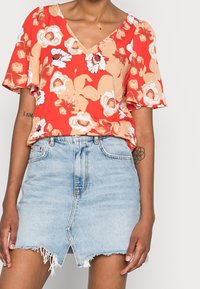 Blusa floral roja con mangas cortas con volantes, combinada con una minifalda de mezclilla azul claro que presenta un dobladillo deshilachado y cremallera frontal.