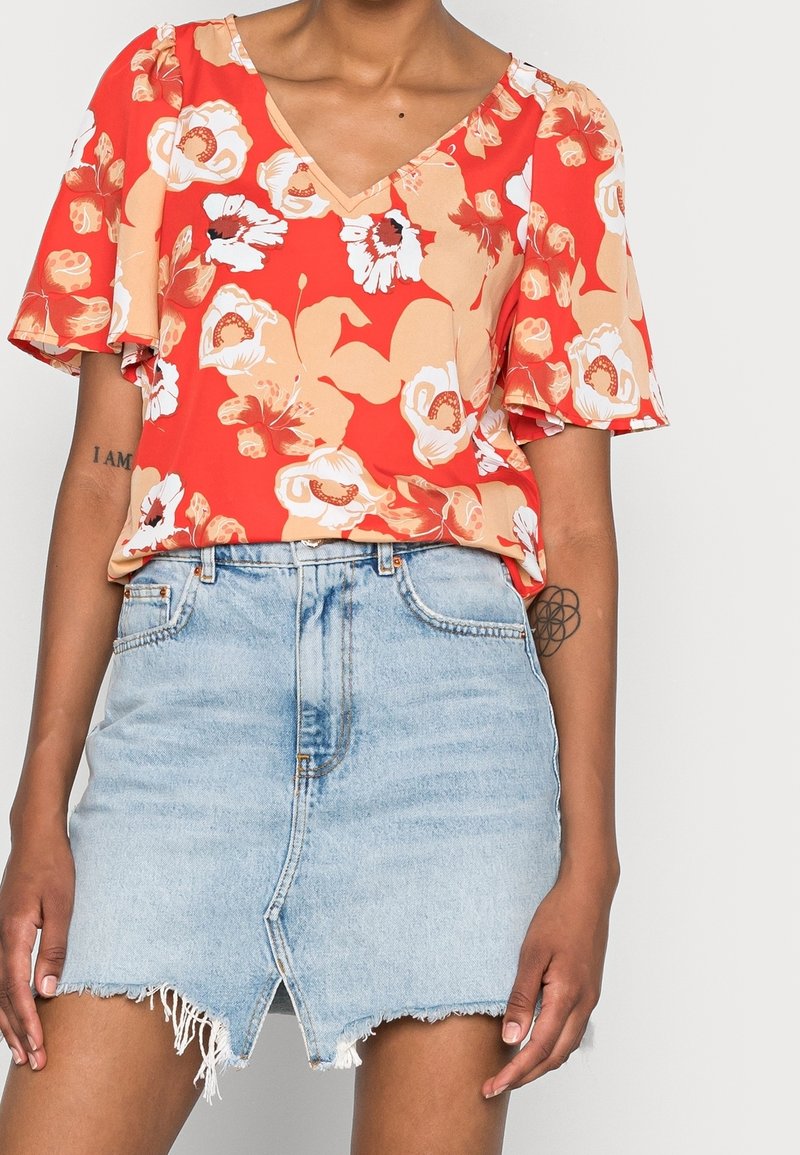 Blusa floral roja con mangas cortas con volantes, combinada con una minifalda de mezclilla azul claro que presenta un dobladillo deshilachado y cremallera frontal.