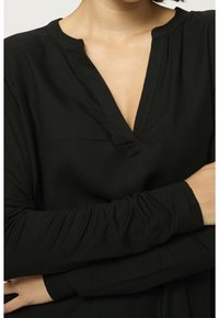 Kvinde iført sort langærmet top med dyb V-udskæring og mandarinhals, armene let krydsede over brystet.