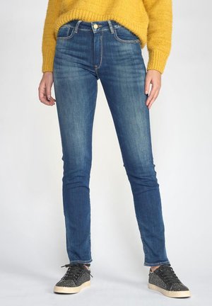 Jeans skinny en denim bleu foncé avec un lavage subtil, présentant un design classique à cinq poches et un bouton doré à la taille. Associés à des baskets noires.