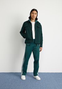 Zelena kordna jakna z gumbom in dvema žepi na prsih, v kombinaciji s turkiznimi hlačami z belimi stranskimi črtami in belimi supergami.