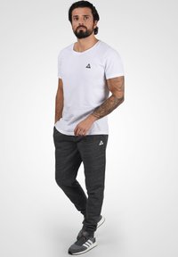 Camiseta blanca de manga corta con un pequeño logo triangular negro, combinada con pantalones jogger grises que presentan una tela texturizada y puños en los tobillos.