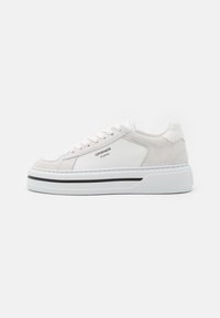 Sneakers blanches en cuir et en daim avec une semelle en caoutchouc texturée, présentant des accents noirs minimalistes et une marque discrète sur le côté.