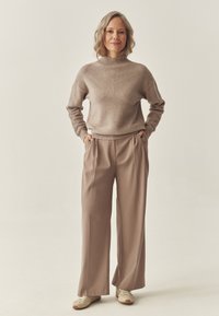 Světle béžový svetr s texturovaným, žebrovaným designem a vysokým výstřihem, doplněný volnými, taupe kalhotami a světlými teniskami.