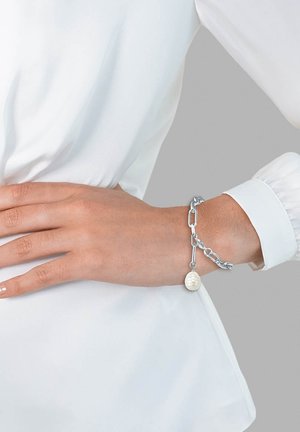 Pulsera de cadena de plata que presenta una perla blanca brillante, combinando eslabones de diferentes tamaños. Llevada en una mano con una manicura neutra.