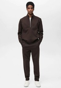 Giacca marrone con zip abbinata a pantaloni, realizzata in un tessuto morbido. Presenta tasche laterali ed è abbinata a sneaker bianche.