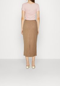 Hnědá žebrovaná midi sukně s rozparkem na zadní straně, spárovaná s světle růžovým cropped topem s vlnkovými okraji. Model má na sobě bílé platformové sandály.