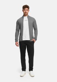 Grå strikket cardigan med glidelås foran og strukturert mønster, båret over en hvit t-skjorte, kombinert med svarte bukser og hvite joggesko.