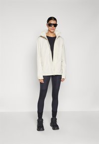 Lys kremfarget vanntett jakke med høy krage, iført over en svart topp og marineblå leggings. Svart sko og solbriller fullfører antrekket.