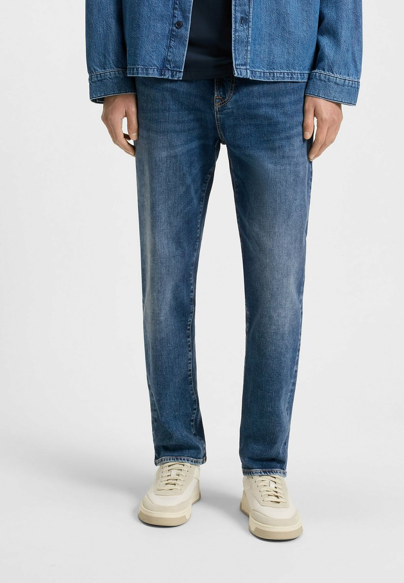 Chaqueta de mezclilla en azul medio, con cierre frontal de botones y dos bolsillos en el pecho. Combinada con jeans de azul claro y zapatillas color crema.