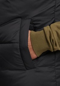 Schwarze gepolsterte Jacke mit einer Seitentasche und einem olivgrünen, gerippten Bündchen. Das Material ist glatt mit sichtbaren Nähdetails.