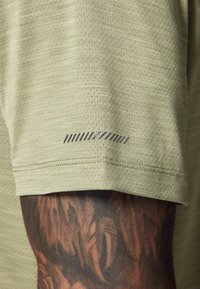 Lys olivenfarvet atletisk tshirt med en struktureret overflade; har en subtil gråstribet accent nær ærmet.