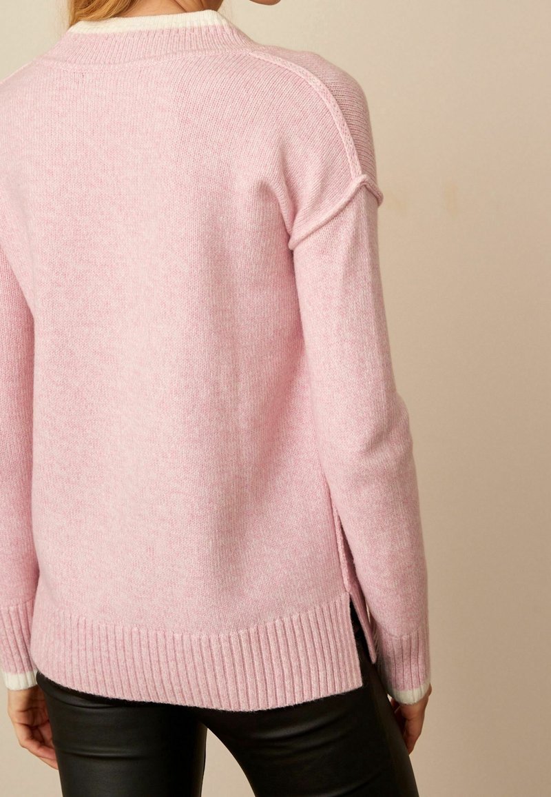 Femme portant un pull rose doux à manches longues avec un ourlet côtelé et des fentes sur les côtés, associé à un pantalon noir, vue de dos.