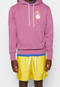 Růžová mikina s klokaní kapsou, bílými šňůrkami a květinovou grafikou. V kombinaci se žlutými šortkami s bílým logem a elastickým pasem.