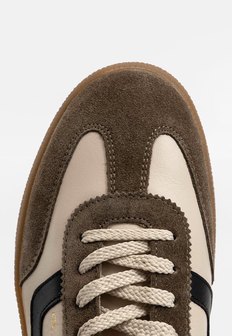 Sneaker z beżową skórzaną cholewką, brązowymi zamszowymi akcentami, czarną boczną paską, grubymi sznurówkami i teksturowaną gumową podeszwą.