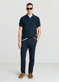 Polo côtelé bleu marine avec accents blancs, associé à un pantalon bleu marine et des chaussures en daim beige. Le modèle porte des lunettes de soleil noires. Fond simple.