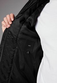 Interiorul unei jachete negre care arată un material neted, buzunar lateral cu eticheta "NEXT" și eticheta de mărime "M" pe un fundal de tricou alb.