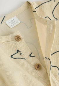 Närbild av en beige babyklädsel med svarta djuransiktstryck, två bruna knappar märkta "NEXT" och en klädlapp "NEXT baby."