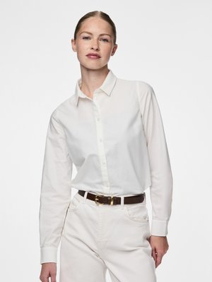 Camisa branca de algodão com botões e mangas compridas, apresentando uma gola clássica e punhos, combinada com calças de cintura alta de cor clara e um cinto castanho.