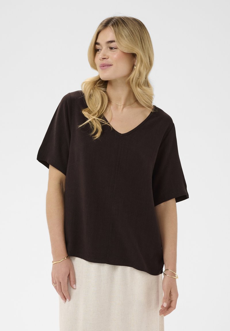 Femme blonde portant un haut ample noir à col en V et manches courtes, ainsi qu'une jupe de couleur claire, debout devant un fond uni.