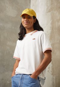 Biała koszulka polo z czerwono-żółtym paskiem przy kołnierzy, z logo na klatce piersiowej, zestawiona z jasnoniebieskimi dżinsami i żółtą czapką. Gładka tekstura materiału.