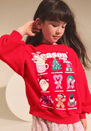 Roter Pullover mit festlichen Illustrationen, darunter eine Tasse heiße Schokolade, ein Weihnachtsbaum und verschiedene Feiertagsleckereien in kontrastierenden Farben.