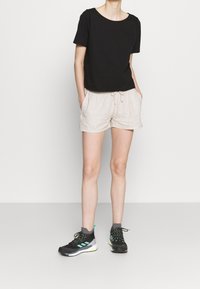 Camiseta negra de manga corta, pantalones cortos beige con cordón y zapatillas deportivas negras con acentos verdes, exhibidos sobre un fondo blanco liso.