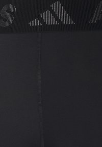 Gros plan sur un tissu noir montrant une couture verticale et une ceinture côtelée avec un logo adidas pixelisé blanc et une inscription partielle.