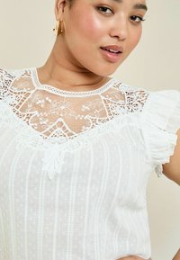 Blusa bianca con dettagli in pizzo trasparente e ricami sul collo. Presenta maniche arricciate e un motivo a righe verticali.