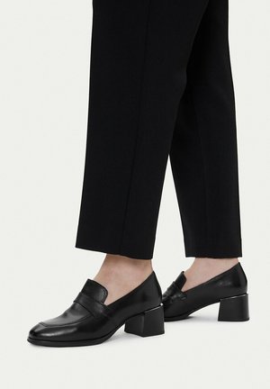 Mocasines de cuero negro con tacón bajo y cuadrado, usados con pantalones negros cortos en pies de piel clara, mostrados sobre un fondo claro.