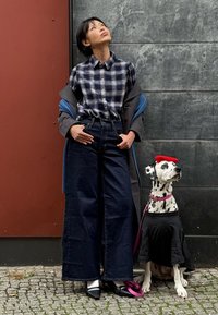 Mulher com camisa xadrez, jeans escuros de perna larga e saltos pretos, parada ao lado de um dálmata vestido com uma camisa preta e boina vermelha.