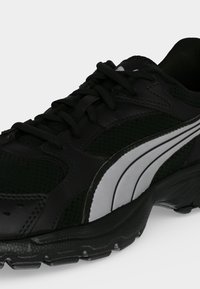 Chaussure de sport noire avec lacets noirs, semelle texturée et un motif de bande blanche courbée sur le côté.